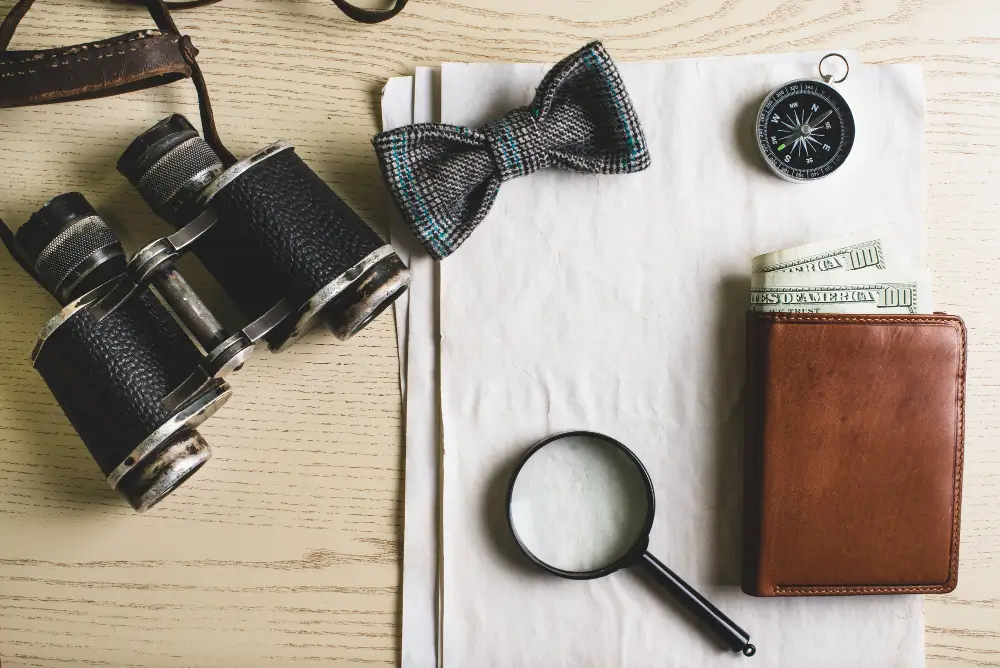 משקפת של חוקר פרטי לצד זכוכית מגדלת ארנק עם שטרות כסף שמציצים משם, פפיון ומצפן